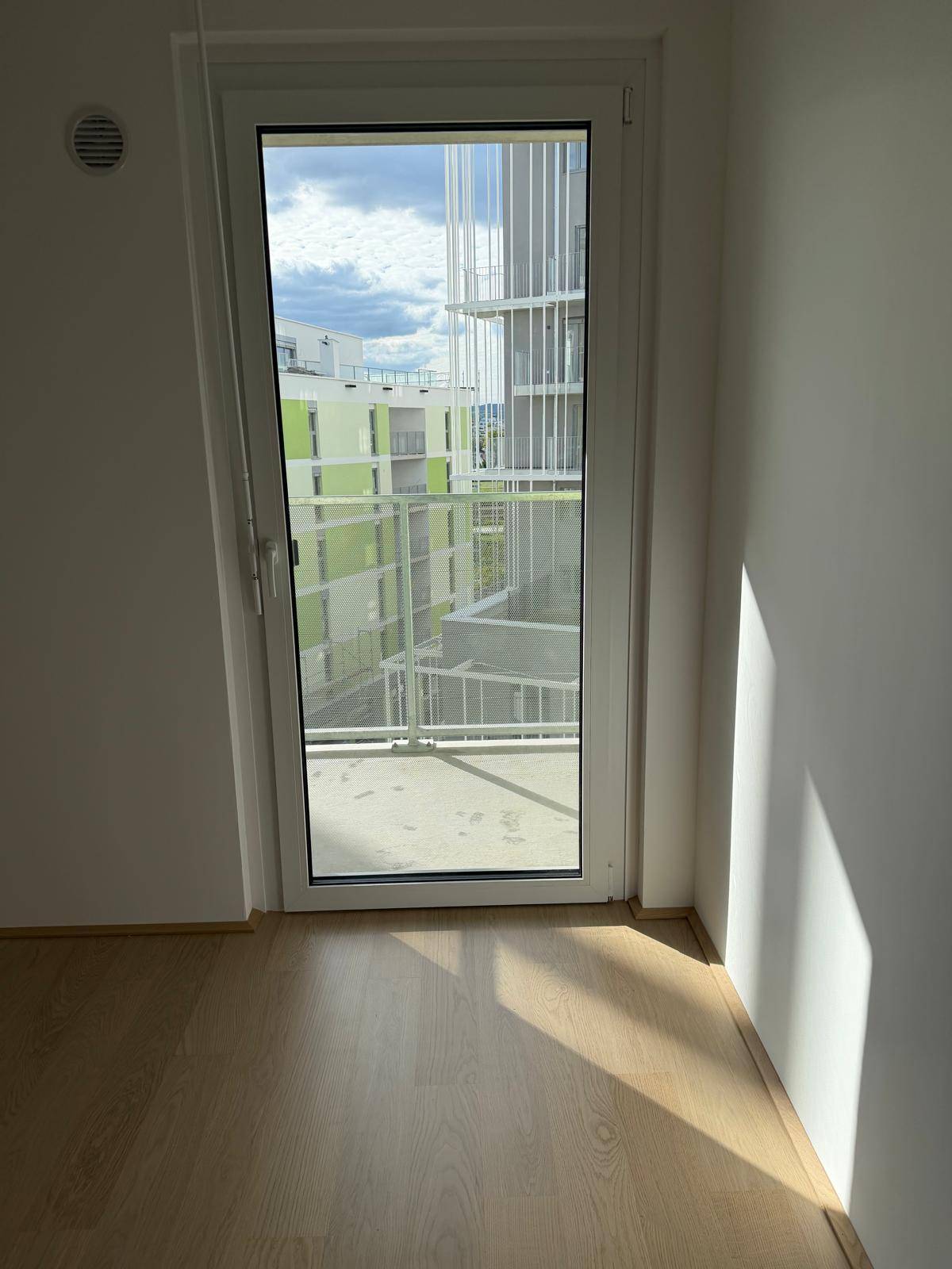 Helles Zimmer mit Holzboden und einer hohen Glastür, die den Blick auf die umliegenden Gebäude freigibt.