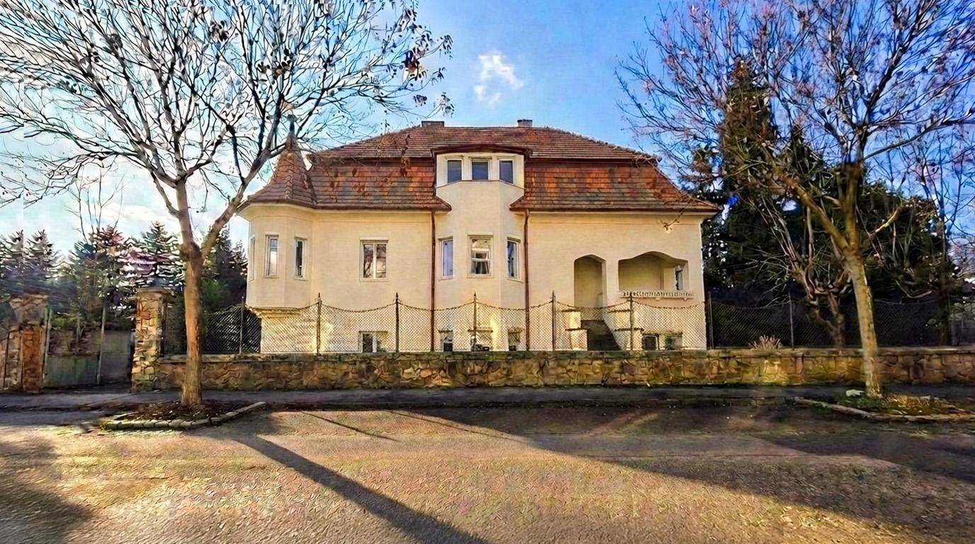 Historische Villa mit markantem Ziegeldach und gepflegtem Vorgarten an einem sonnigen Tag.