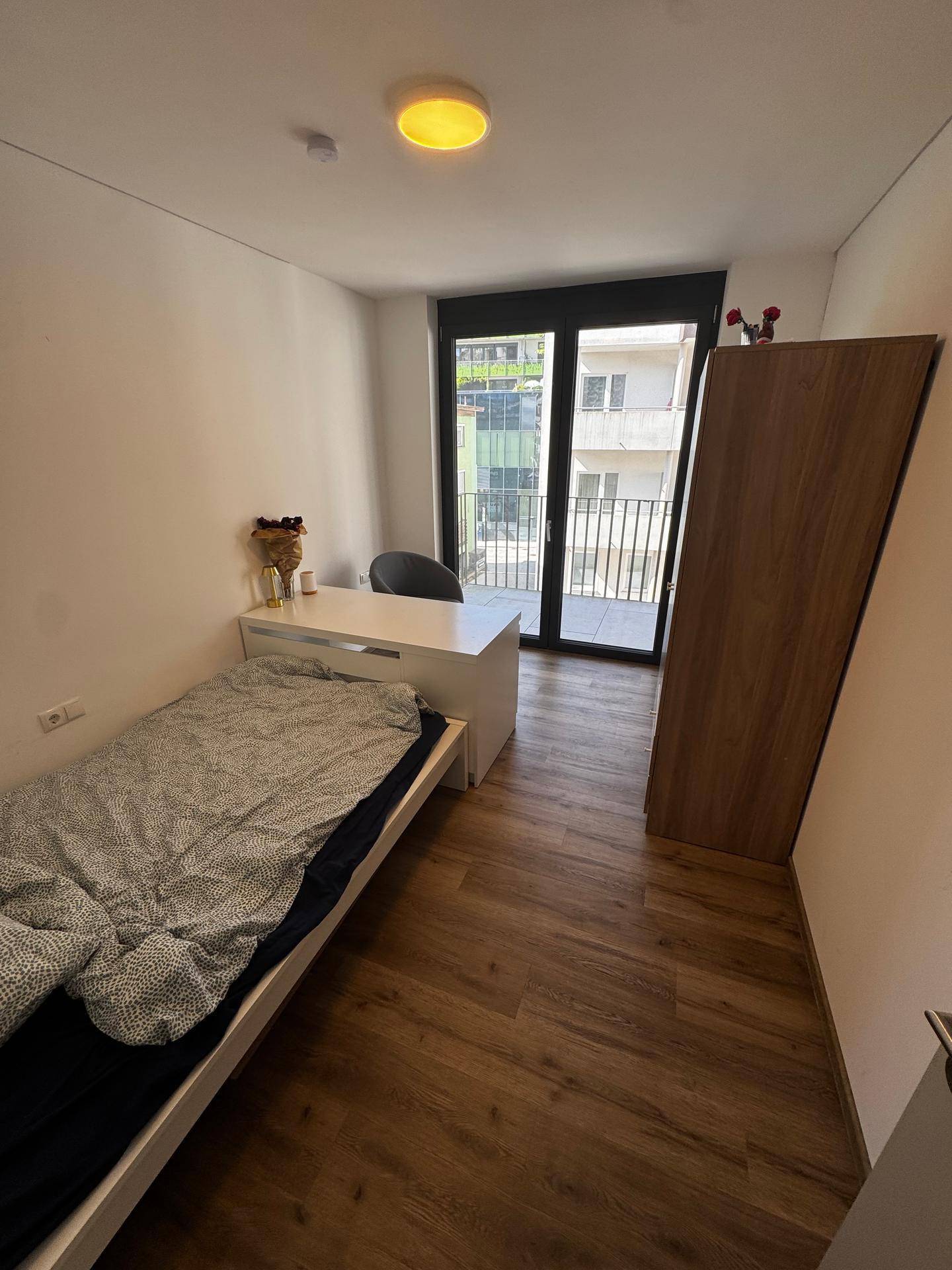 Schlafzimmer mit Holzboden, Einzelbett, Schreibtisch und einem großen Kleiderschrank neben dem Balkonzugang.