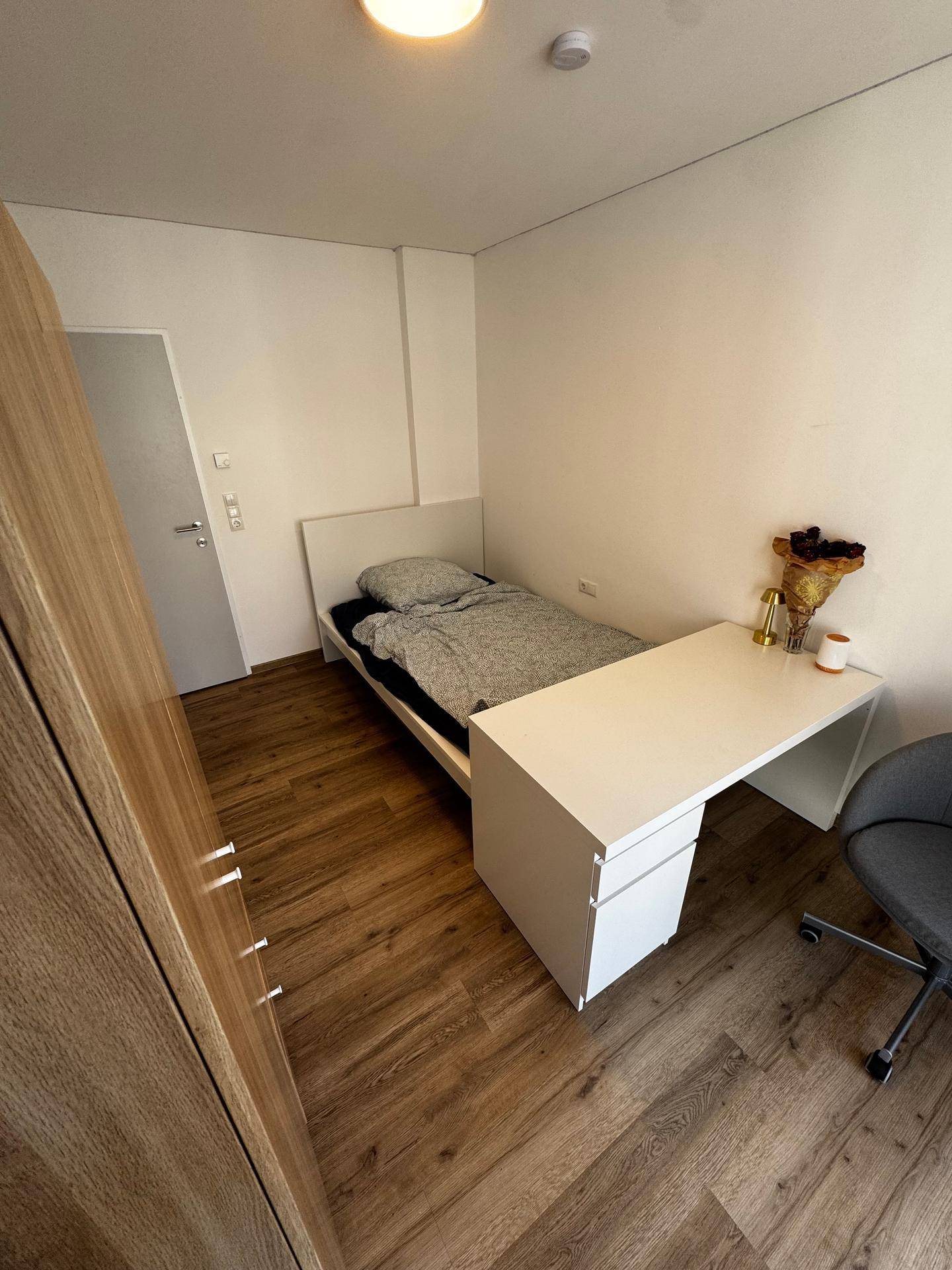 Schlafzimmer mit Holzboden, Einzelbett, Schreibtisch und einem großen Kleiderschrank nahe der Zimmertür.