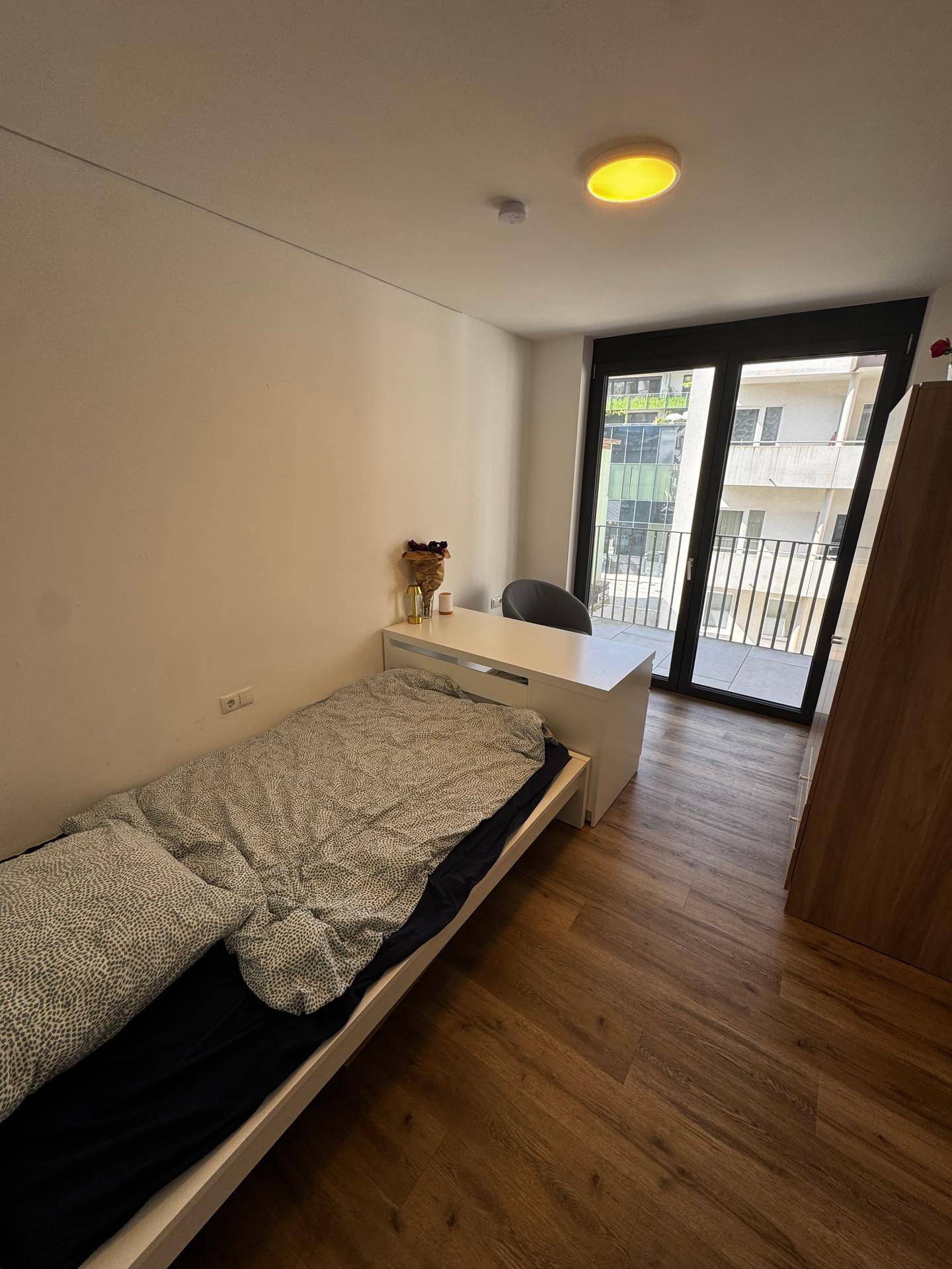 Geräumiges Schlafzimmer mit Holzboden, Einzelbett, Schreibtisch und großem Fenster zum Balkon.