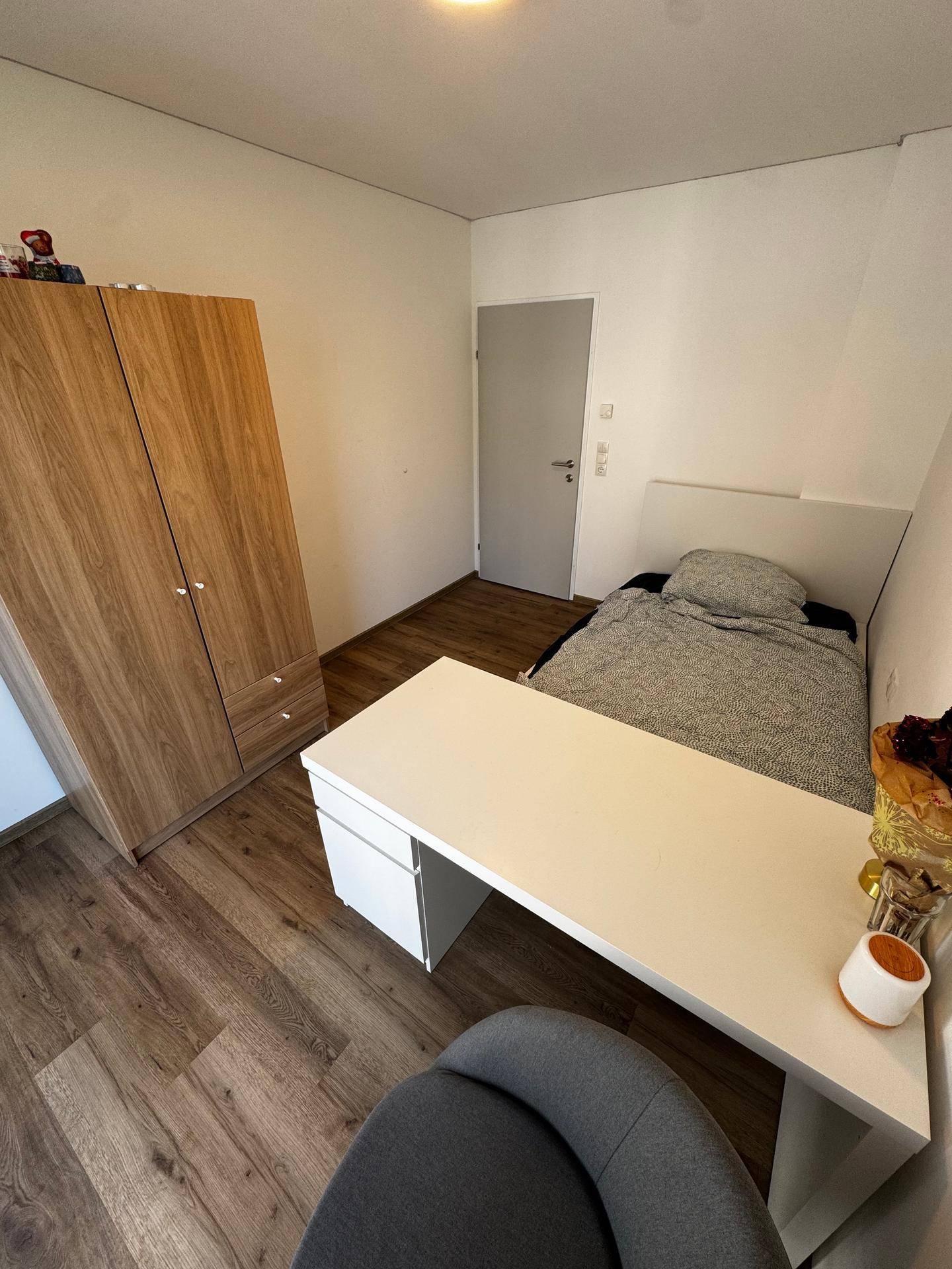 Heller Raum mit Holzboden, Einzelbett, Schreibtisch und einem geräumigen Kleiderschrank.