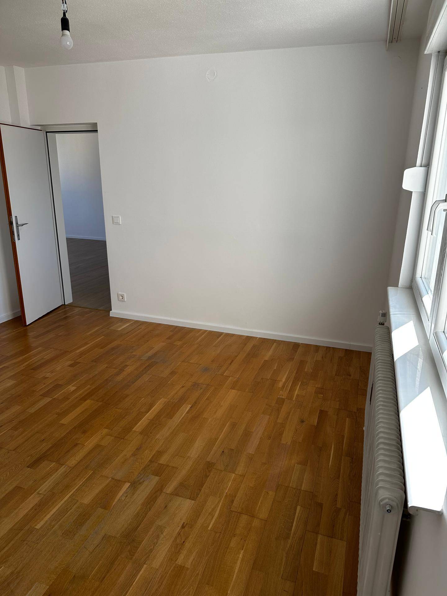 Helles Zimmer mit klassischem Parkettboden, weißen Wänden und großem Fenster.