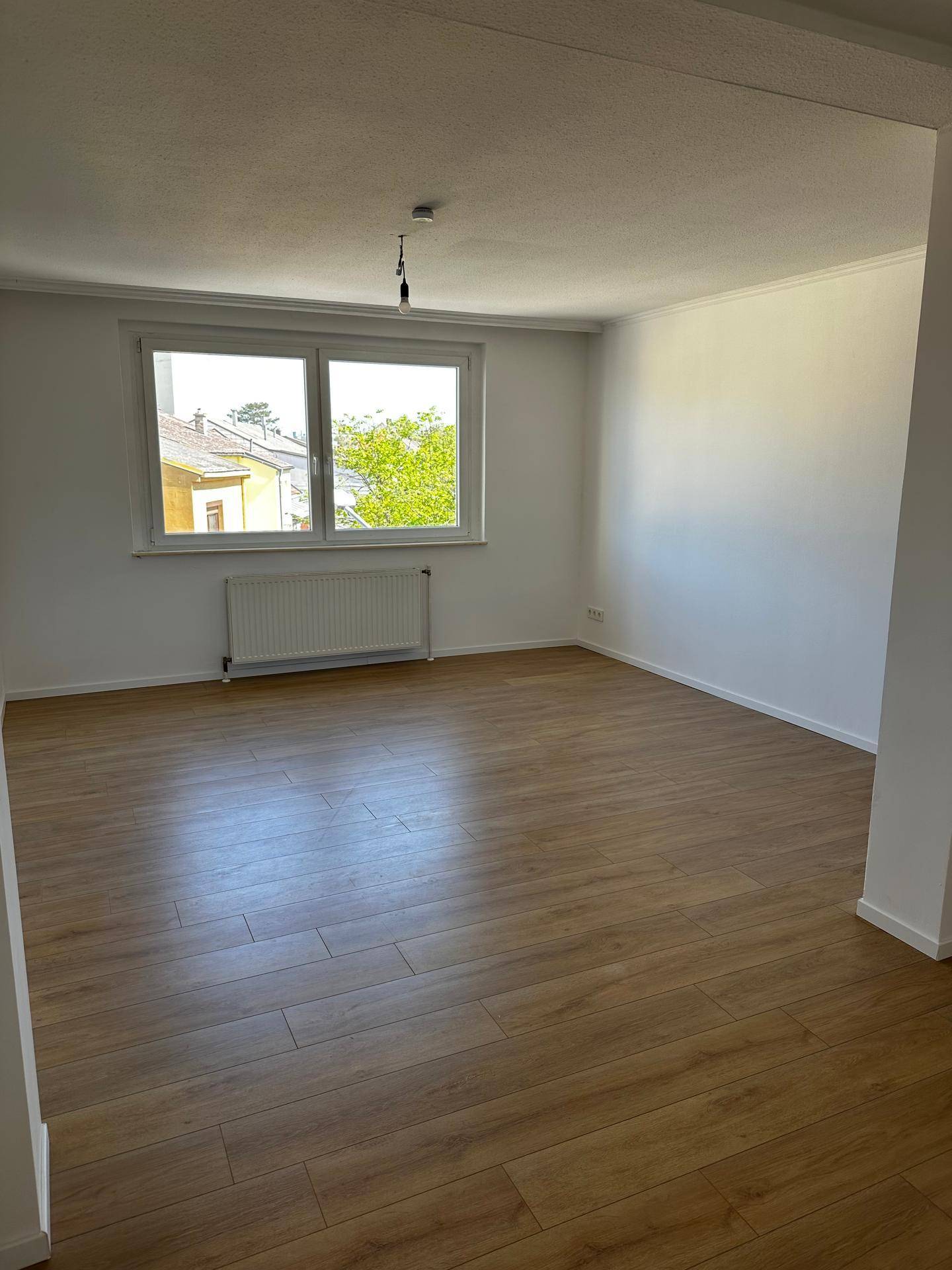 Geräumiger, heller Wohnbereich mit Laminatboden, großen Fenstern und Heizkörper unter dem Fenster.