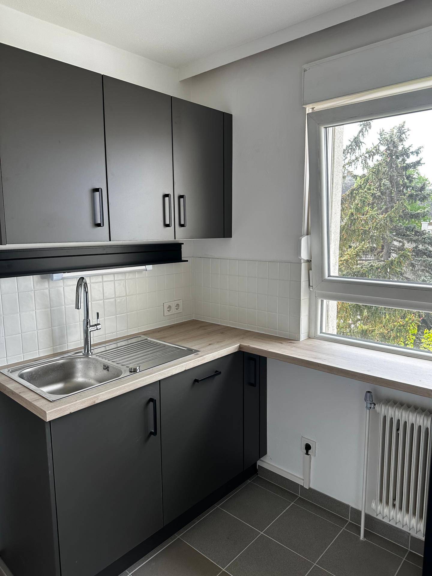 Helle Küche mit Spülbecken, dunklen Unterschränken, Holzoptik-Arbeitsplatte und Fenster mit Blick ins Grüne.