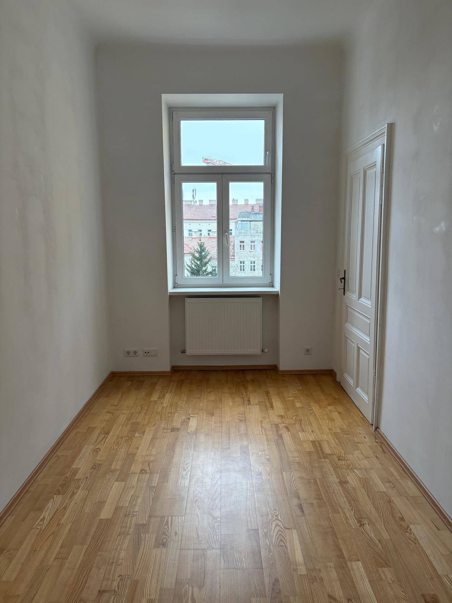 Leerer Raum mit hellem Parkettboden und großem Fenster, das viel Licht hereinlässt.
