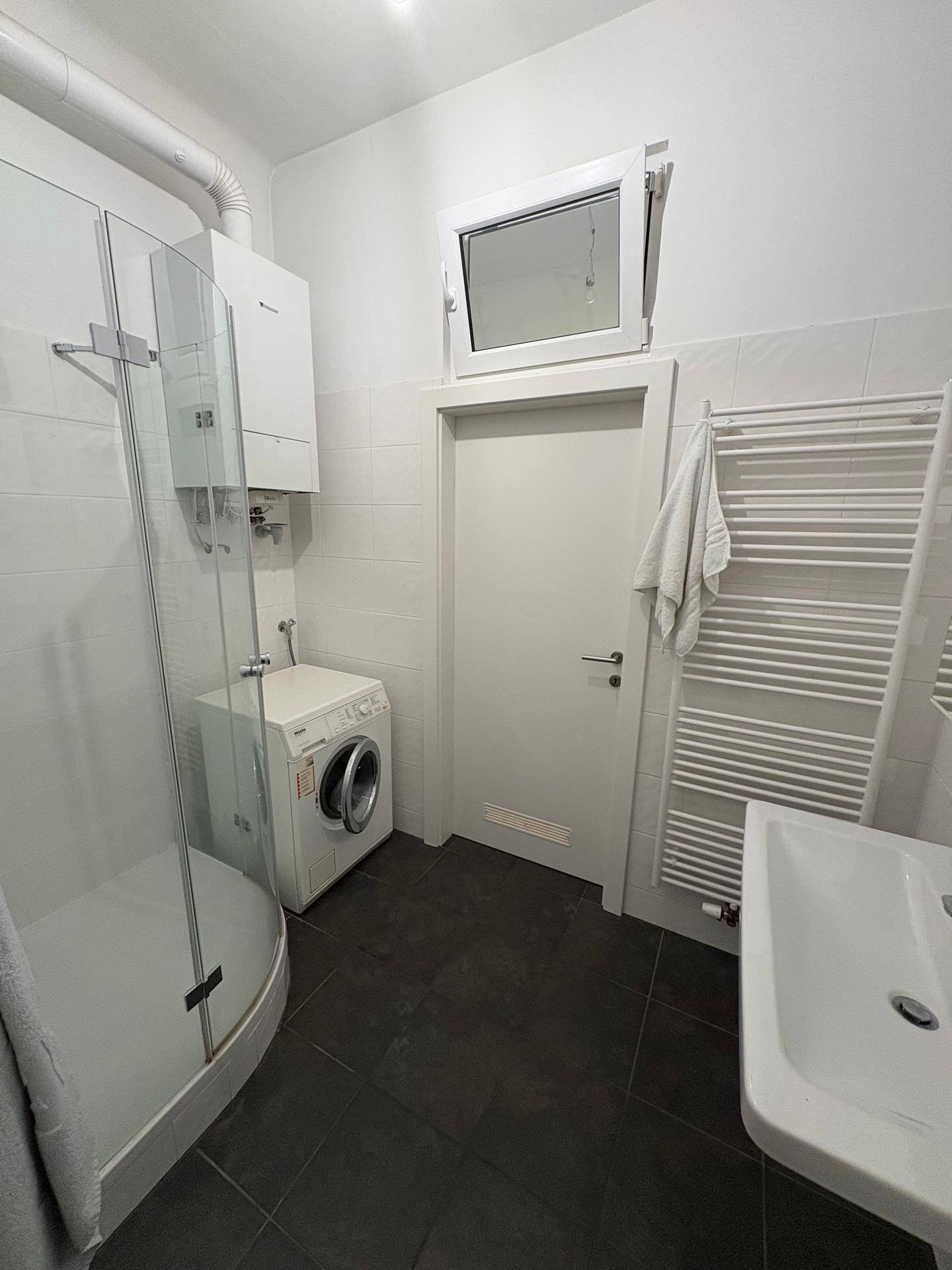 Helles Badezimmer mit Dusche, Waschmaschine, Handtuchheizkörper und kleinem Fenster.