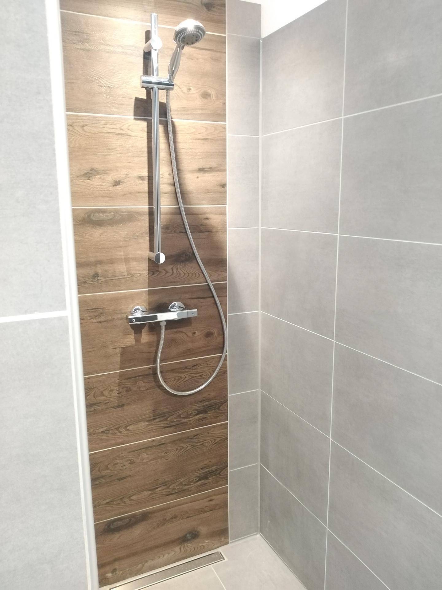 Moderne Duschkabine mit grauen Fliesen und einer Akzentwand in Holzoptik.