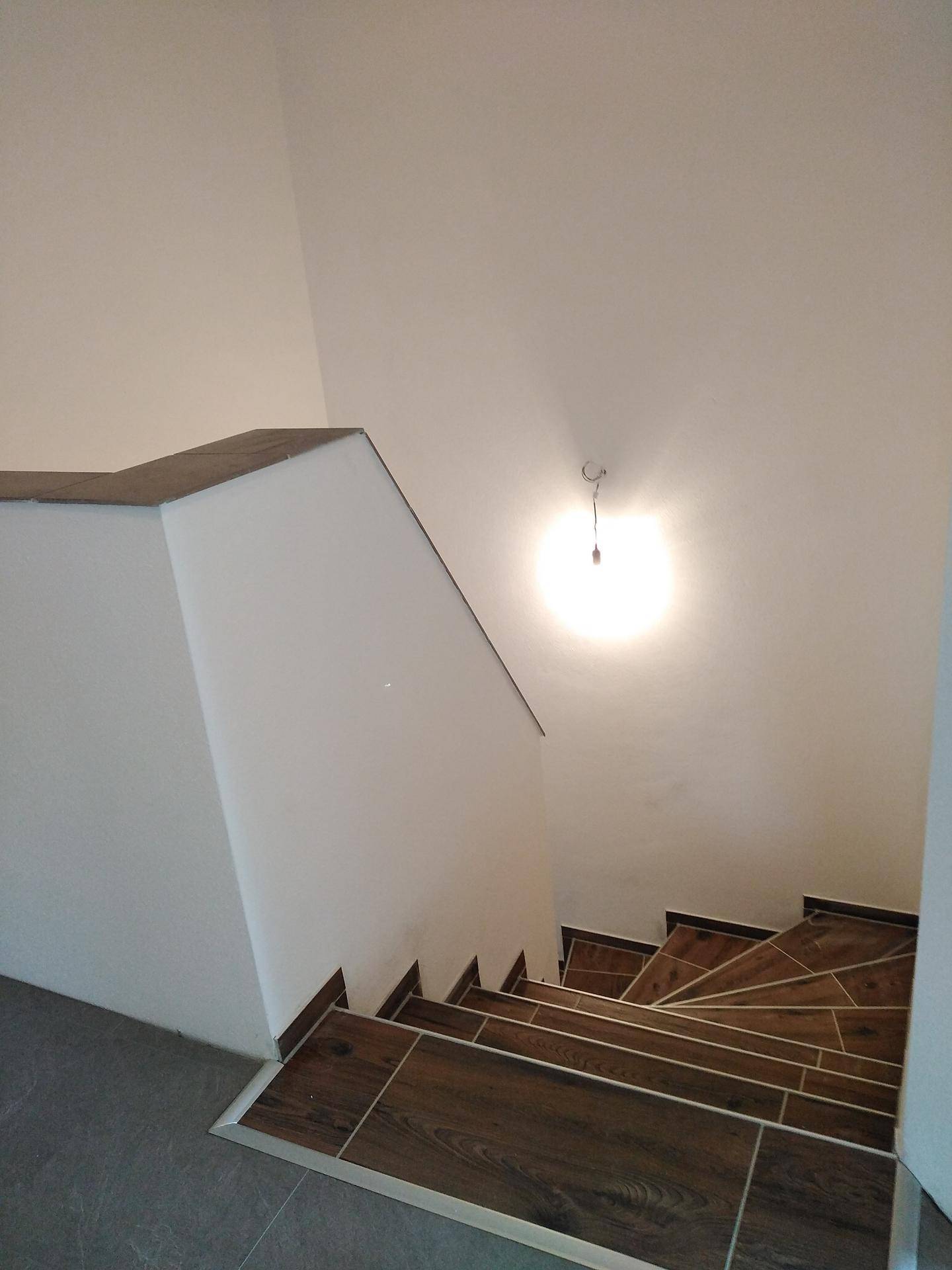Moderner Treppenaufgang mit Holzoptikfliesen und weißen Wänden, beleuchtet von einer einfachen Lampe.