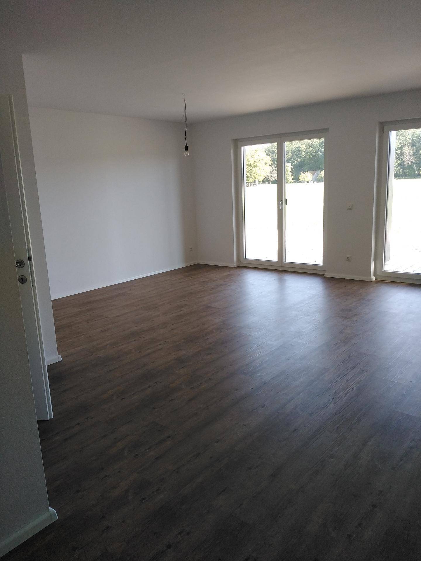 Geräumiges, leeres Wohnzimmer mit dunklem Laminatboden und großen Fenstern, die viel Licht hereinlassen.