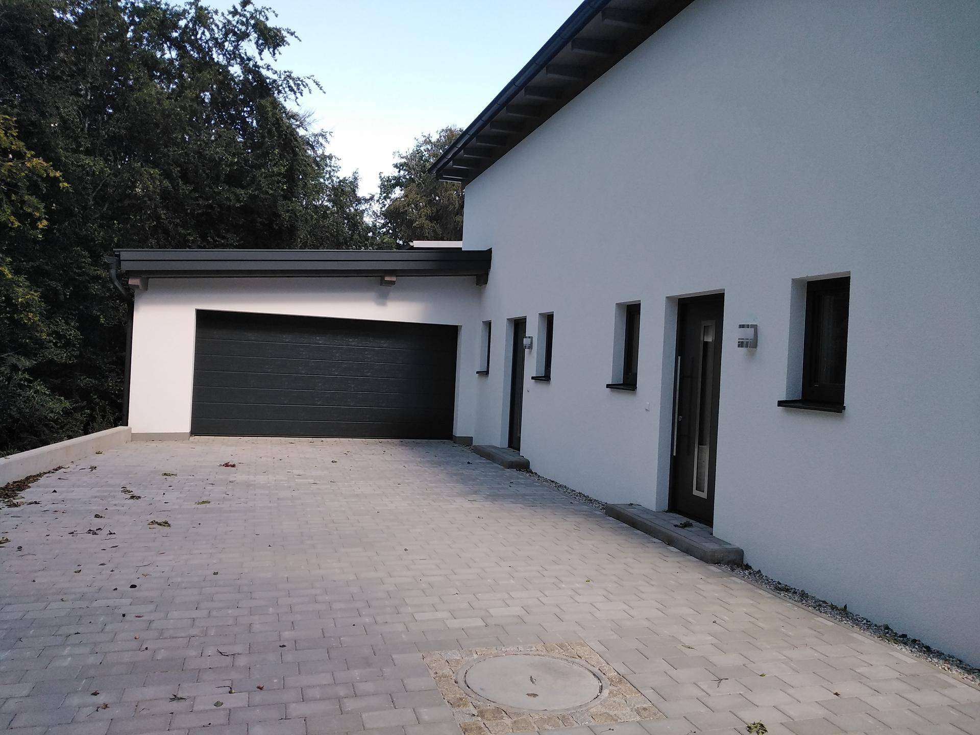 Moderne Hausfassade mit weißem Putz, dunklem Garagentor und gepflasterter Zufahrt.