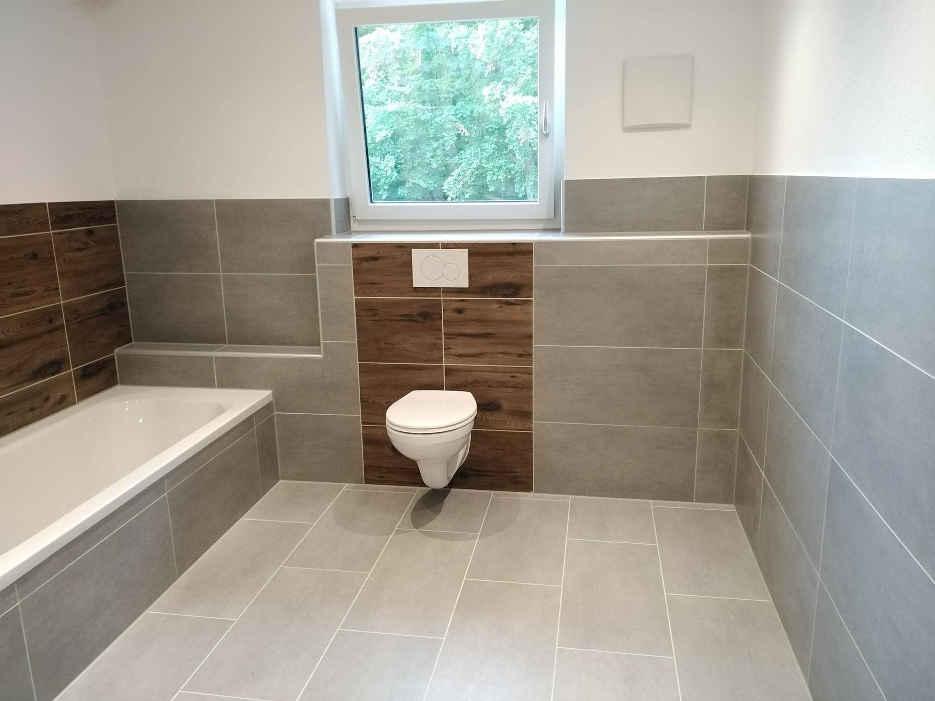 Modernes Badezimmer mit Badewanne, wandhängendem WC und Fenster, gefliest in Grau und Holzoptik.