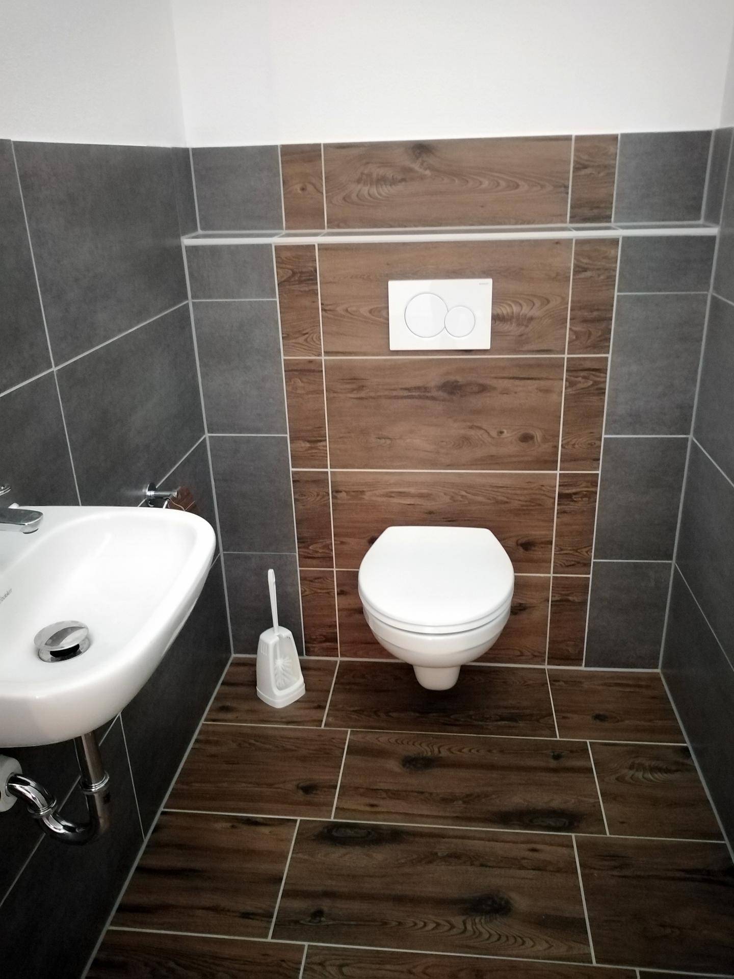 Gäste-WC mit wandhängendem WC und Einzelwaschbecken, gefliest in Grau und Holzoptik.
