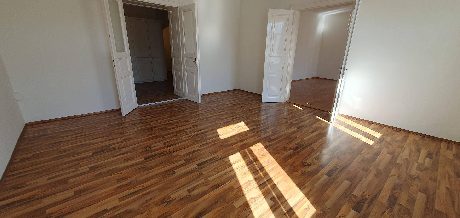 Geräumiger Raum mit glänzendem Holzboden und mehreren Türen, durch die Sonnenlicht fällt.