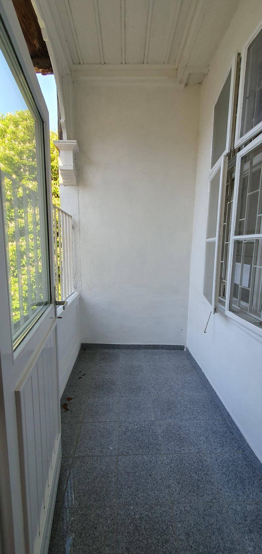 Kleiner Balkon mit grauem Fliesenboden und weißem Geländer, umgeben von Bäumen.