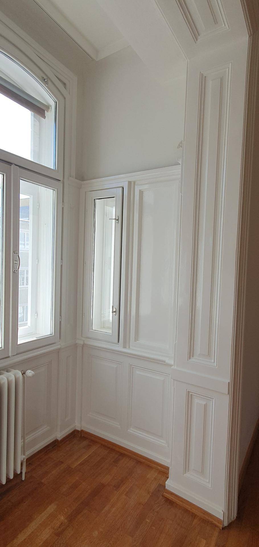Ecke eines Raumes mit hohen Fenstern, Holzvertäfelung und klassischer Stuckverzierung an der Decke.