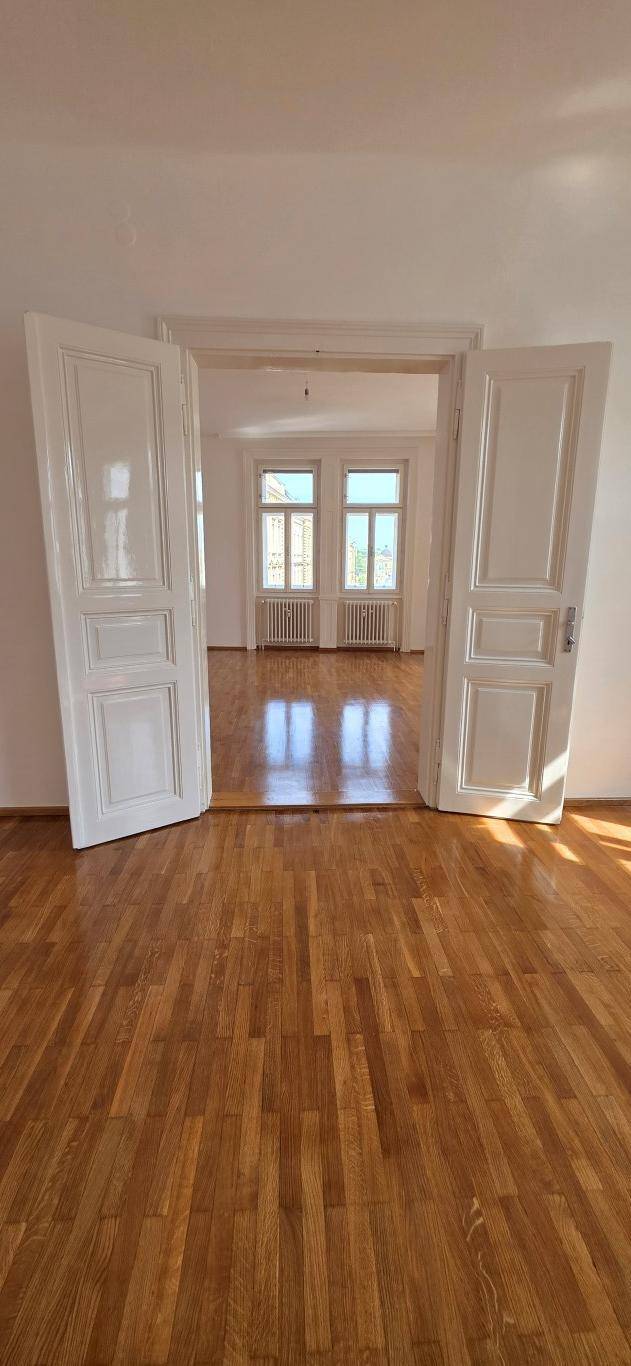 Heller Raum mit Holzboden und Doppeltüren, die den Blick auf weitere Fenster freigeben.