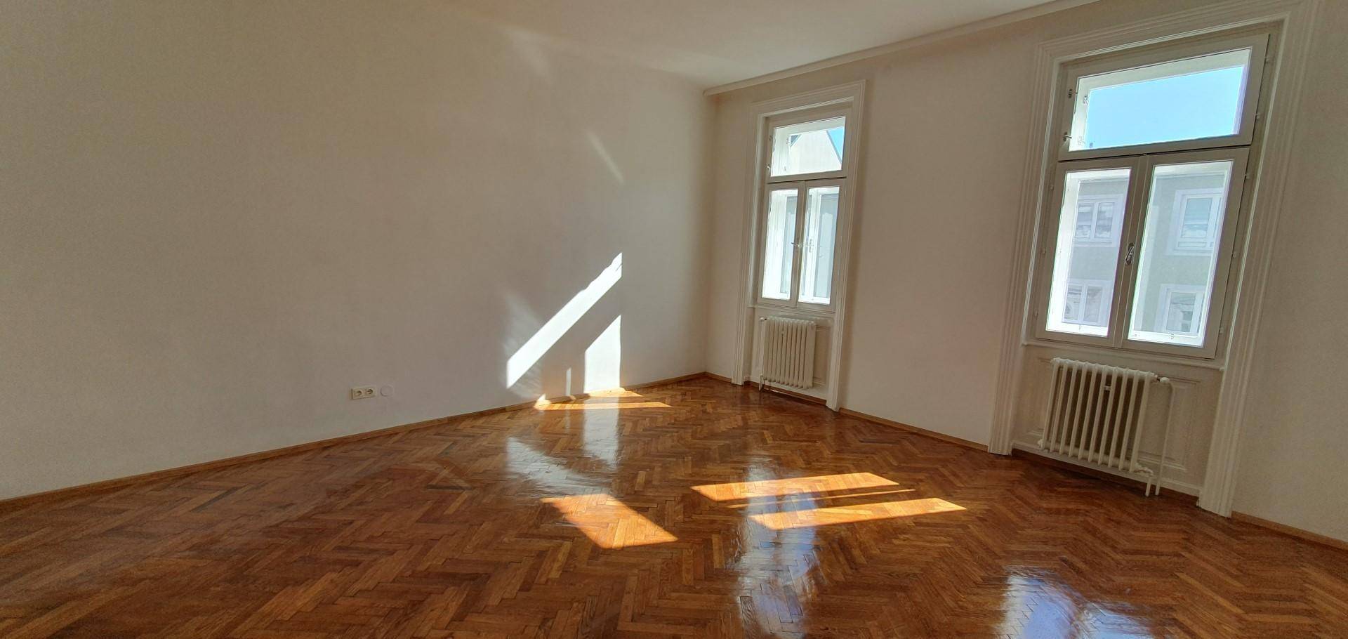Helles Schlafzimmer mit Fischgrätparkett und zwei Fenstern, die den Raum mit Licht füllen.