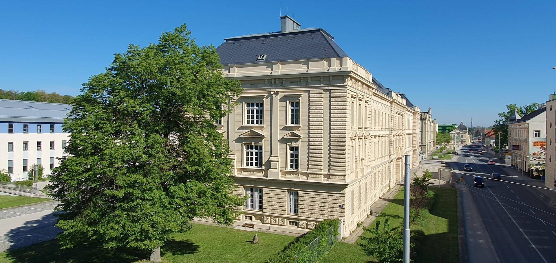 Klassisches Gebäude mit beiger Fassade und vielen Fenstern, umgeben von Grünflächen und einer Straße.
