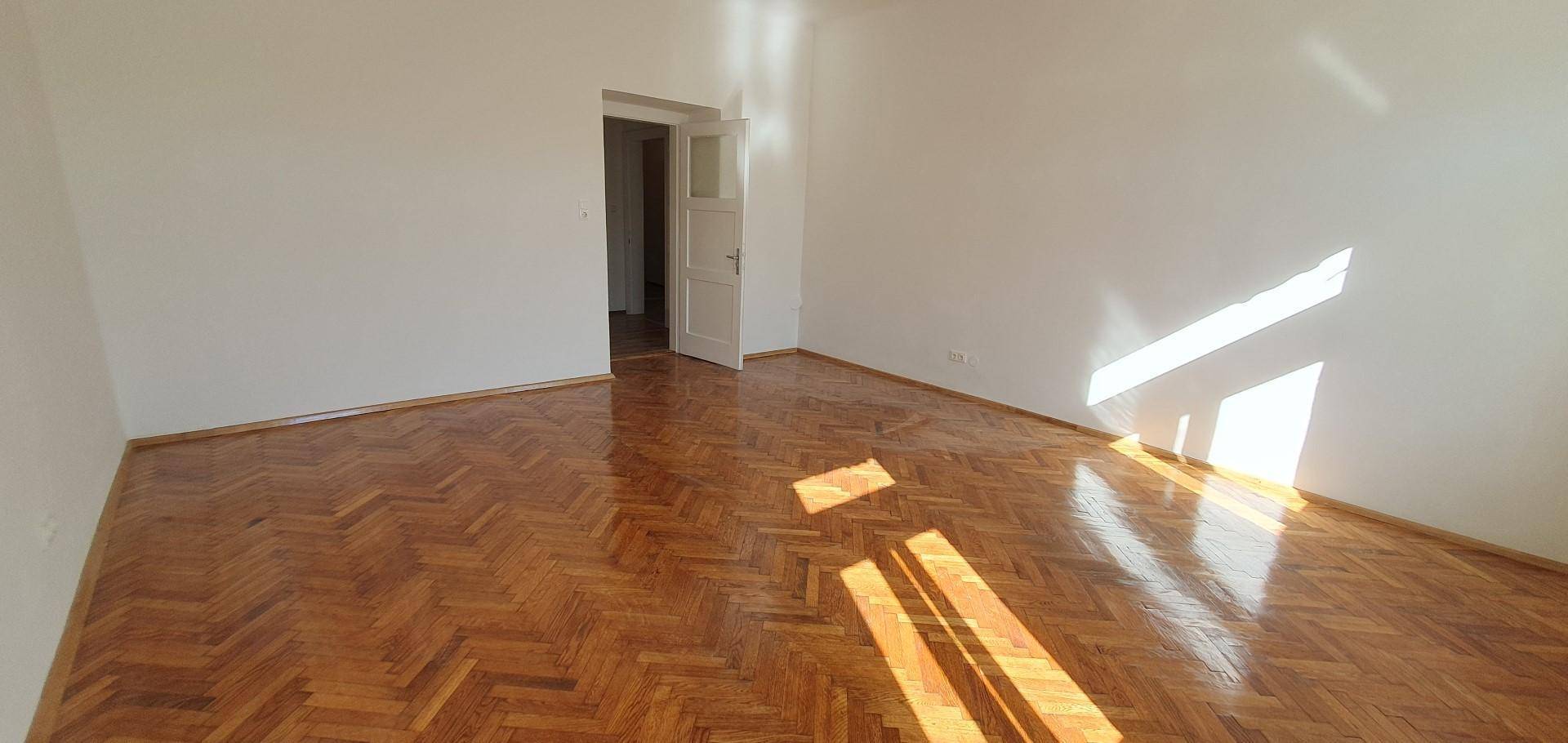 Geräumiges Schlafzimmer mit Fischgrätparkett und einer Tür, durch die Sonnenlicht fällt.