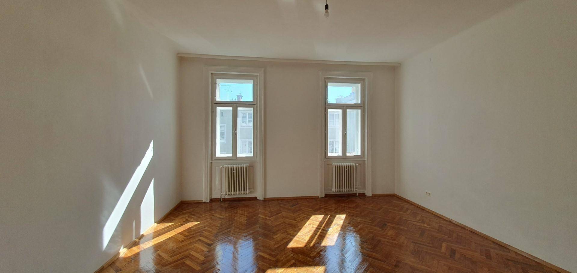 Helles Schlafzimmer mit Fischgrätparkett und zwei Fenstern, durch die Sonnenlicht fällt.