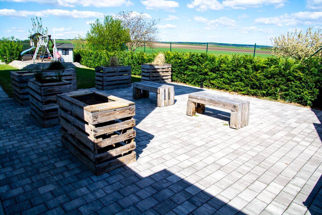 Gepflasterter Terrassenbereich mit Holzbänken und Pflanzgefäßen aus Paletten, umgeben von einer Hecke.