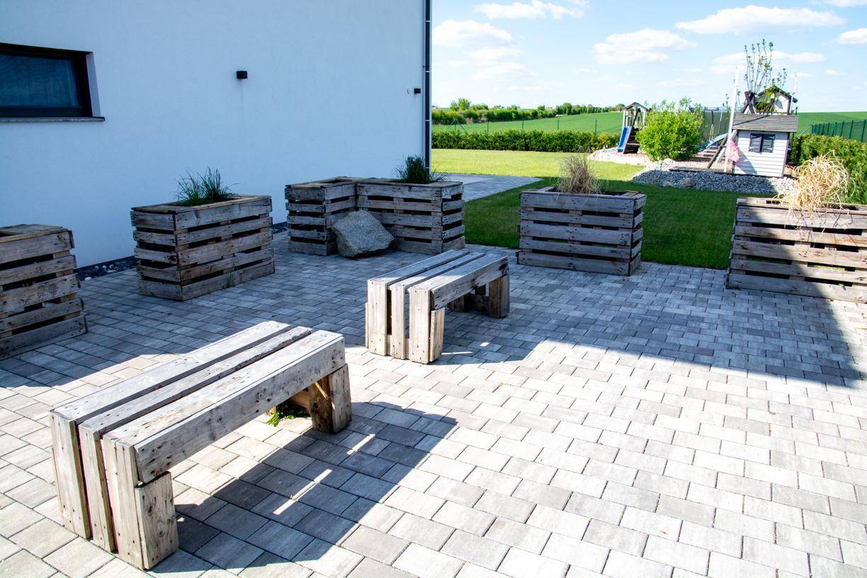Weitere Ansicht des gepflasterten Terrassenbereichs mit Holzbänken und Pflanzgefäßen am Haus.