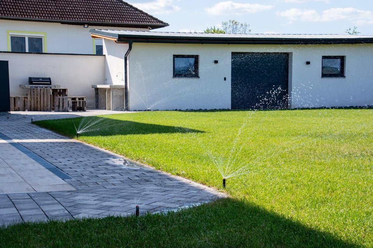 Grüner Rasen mit aktiven Sprinklern, angrenzend an einen gepflasterten Weg und das Haus mit Garage.
