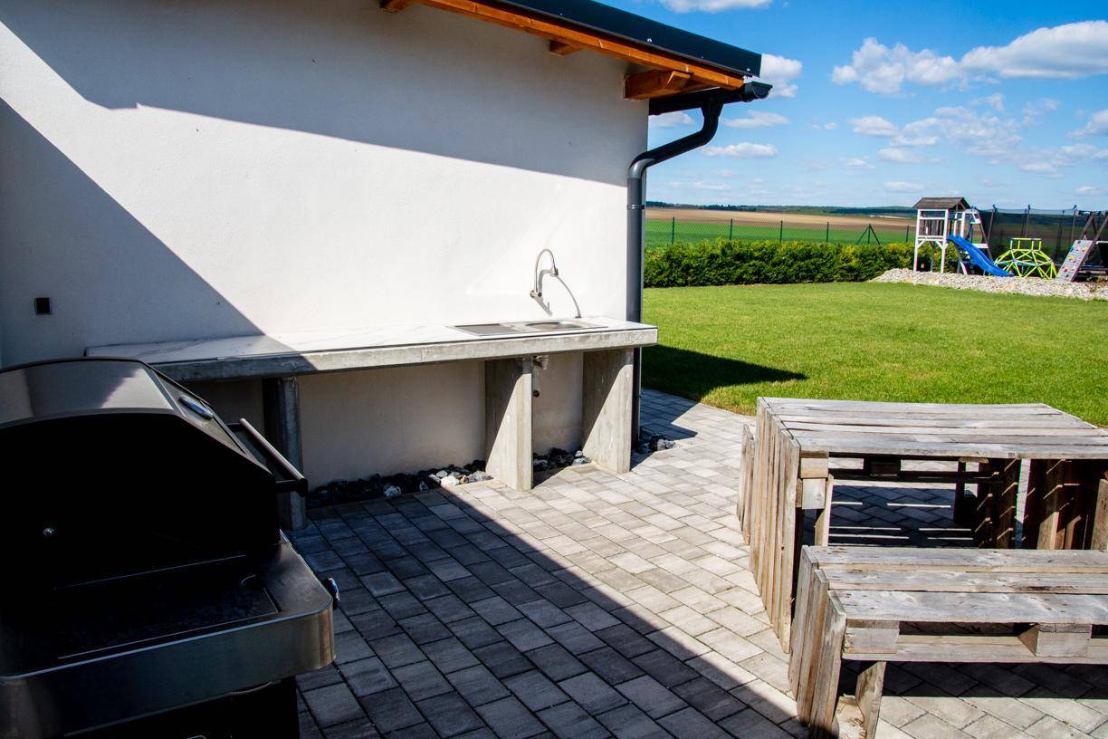 Außenbereich mit Grill und gemauerter Arbeitsplatte, angrenzend an einen gepflasterten Weg und Rasen.