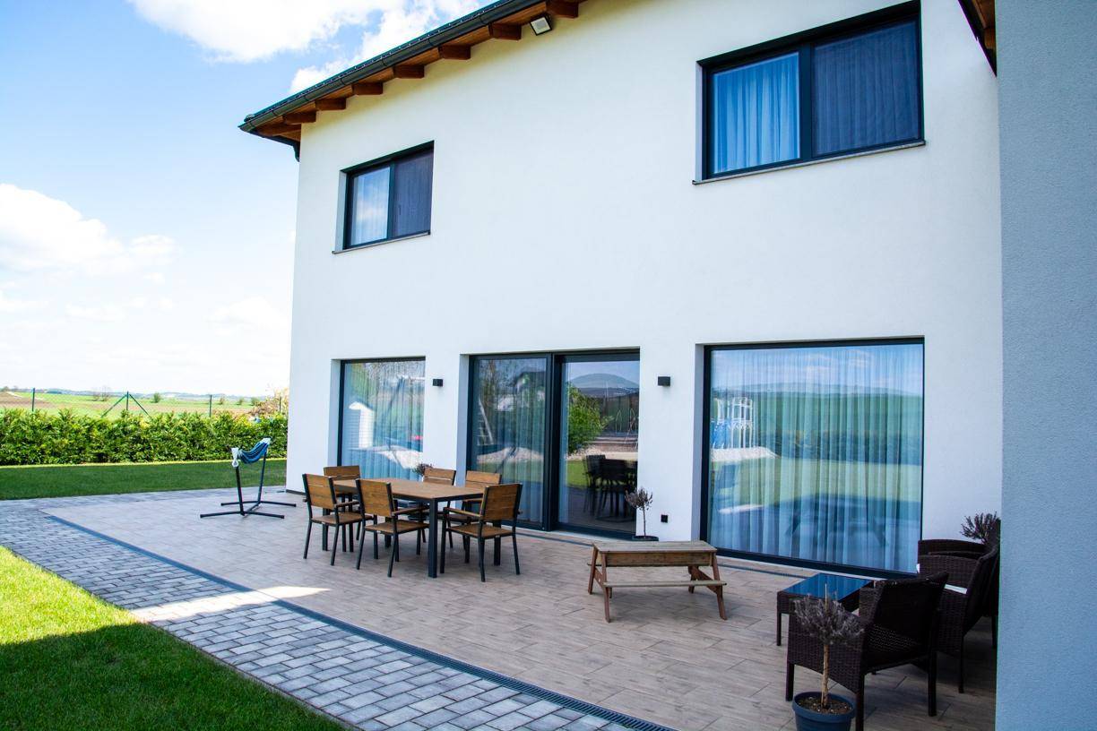 Seitenansicht des Hauses mit Terrasse, Gartenmöbeln und gepflegtem Rasen unter blauem Himmel.