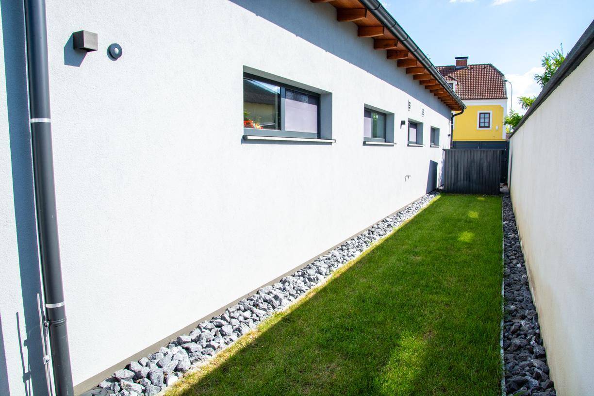 Schmaler Seitengarten mit Rasenfläche und dekorativen Steinen entlang der Hauswand.