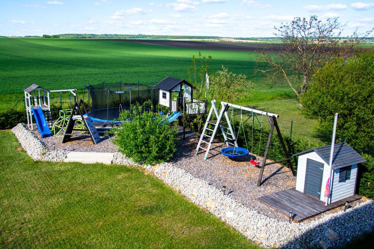 Großer Spielplatz im Garten mit Trampolin, Schaukeln und Rutschen, umgeben von grünen Feldern.