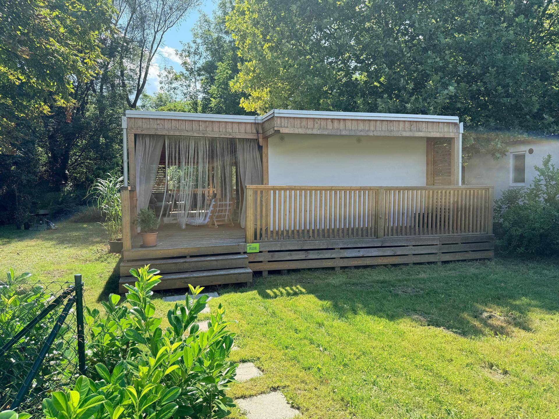 Vorderansicht des Holzchalets mit überdachter Terrasse, Vorhängen und einem gepflegten Rasenbereich.
