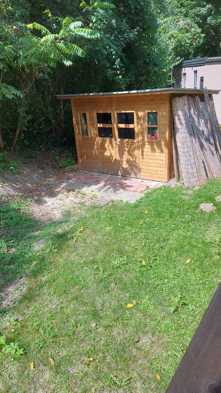 Kleiner Holzschuppen im Garten, umgeben von Rasen und Bäumen.