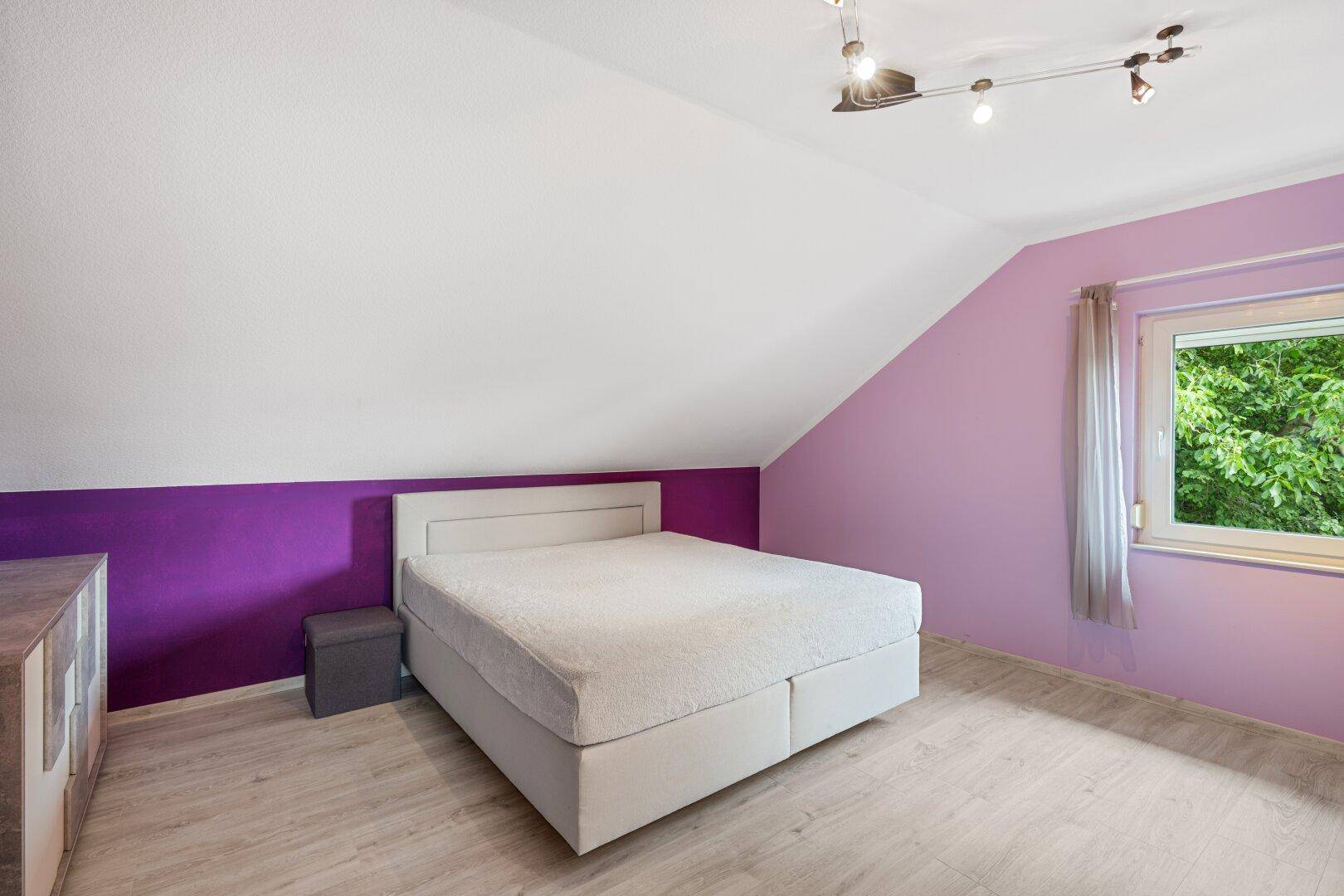Schlafzimmer im Dachgeschoss mit Bett, lila Wänden und Schrägdach.