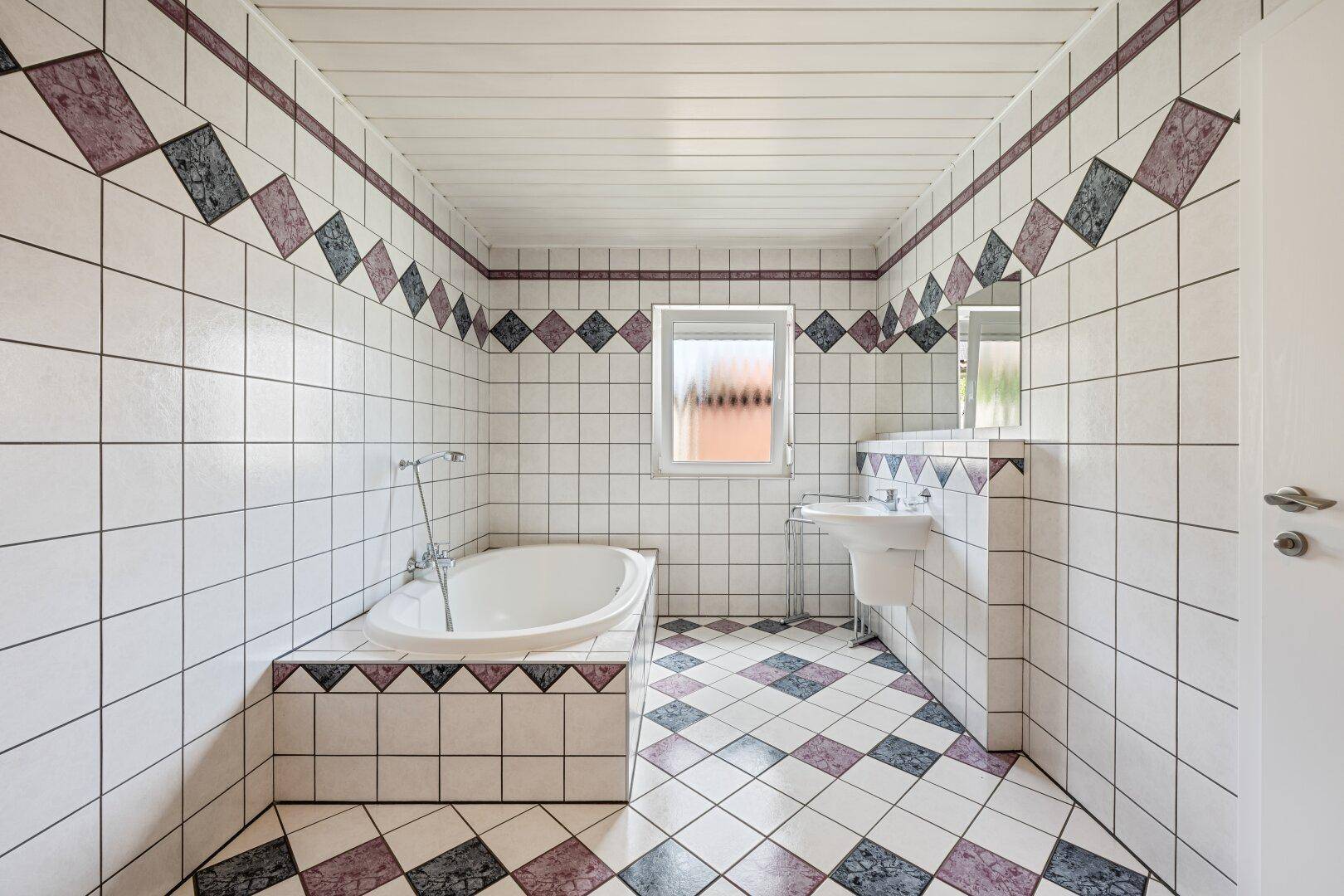 Badezimmer mit Badewanne, Waschbecken und auffälligen Fliesen im 80er-Jahre-Stil.