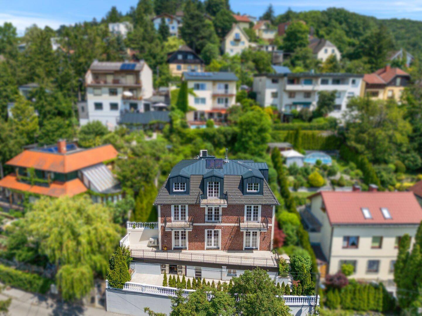 Weitläufige Ansicht des modernen Wohnhauses mit Backsteinfassade, umgeben von viel Grün und weiteren Häusern am Hang.