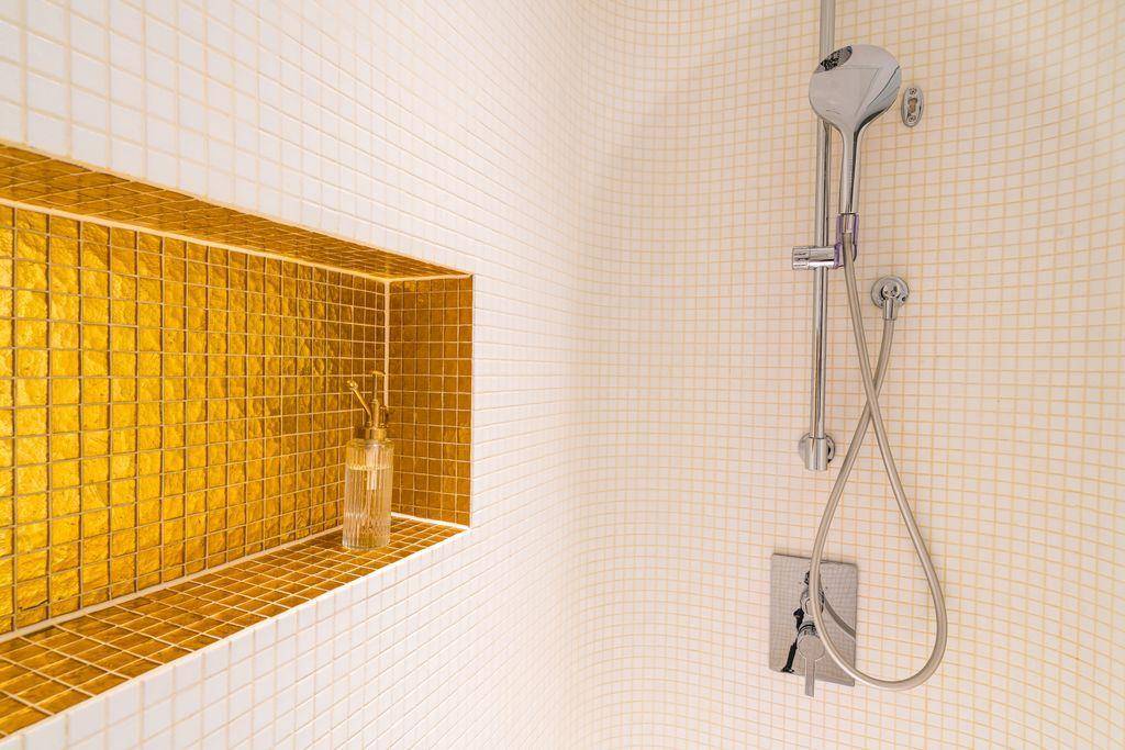 Duschbereich mit eleganten weißen und goldenen Mosaikfliesen und integrierter Ablage.