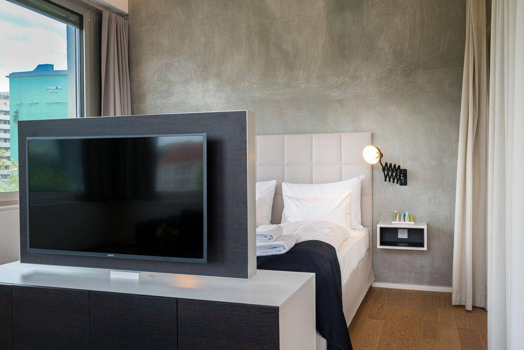 Schlafbereich mit modernem Bett, Fernseher auf einem Sideboard und einer strukturierten grauen Wand.