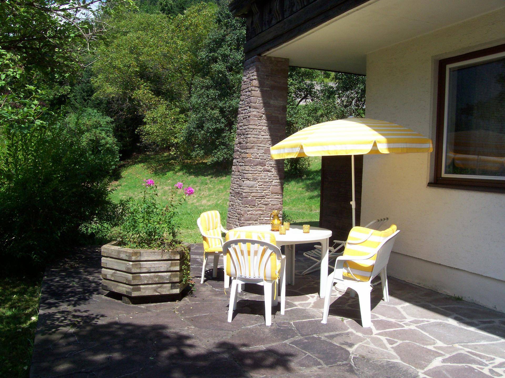 Sonniger Terrassenbereich mit Gartenmöbeln und Sonnenschirm, umgeben von üppigem Grün.