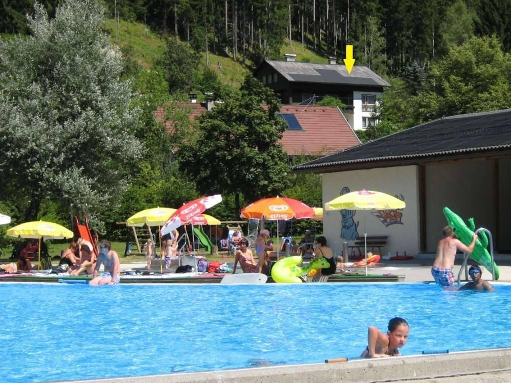 Außenpoolbereich mit Badegästen, Sonnenschirmen und einem Haus im Hintergrund mit Solaranlage.