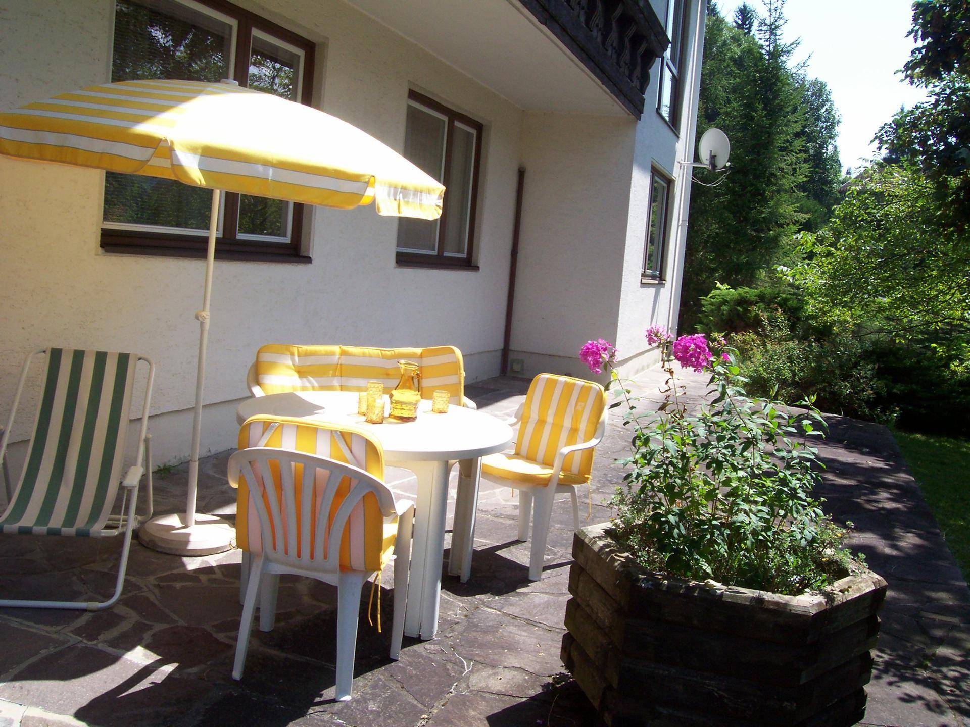 Gemütliche Terrasse mit gelb-weiß gestreiften Gartenmöbeln und Sonnenschirm, umgeben von Grün.