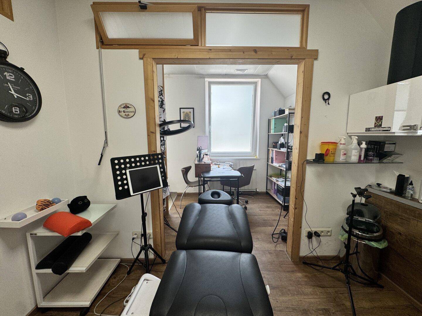 Behandlungsraum mit einer Liege, einem Schreibtisch und Regalen, verbunden mit einem weiteren Arbeitsbereich.