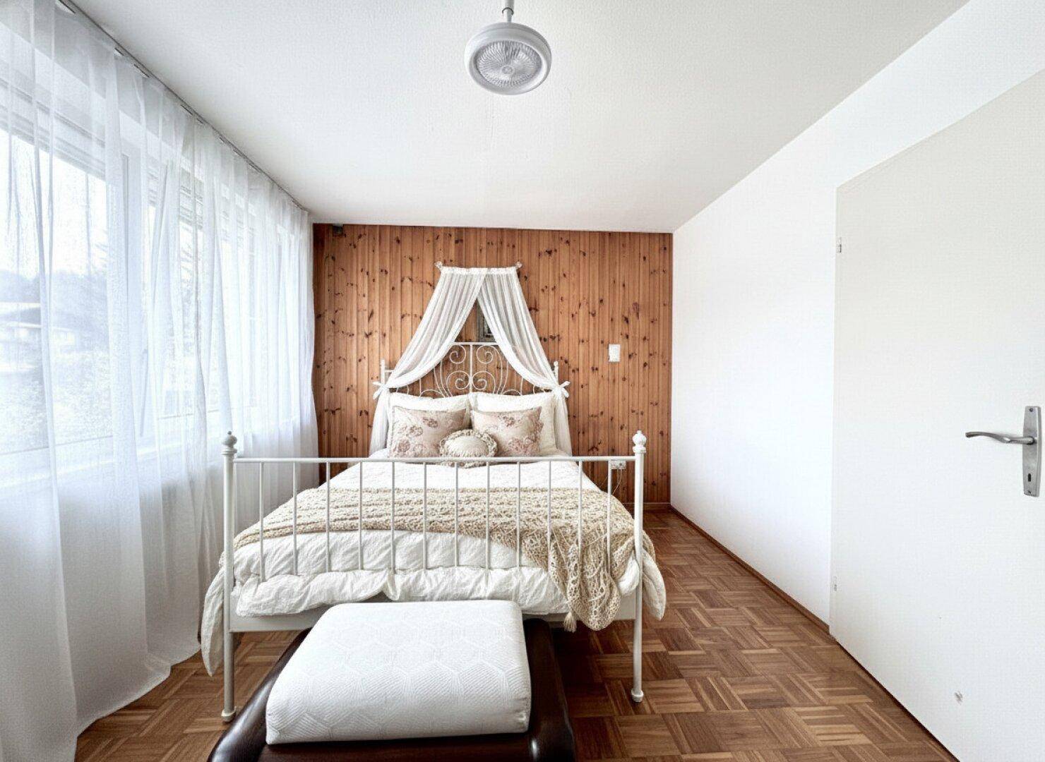 Gemütliches Schlafzimmer mit Holzwand, Parkettboden und einem Himmelbett unter einem Deckenventilator.