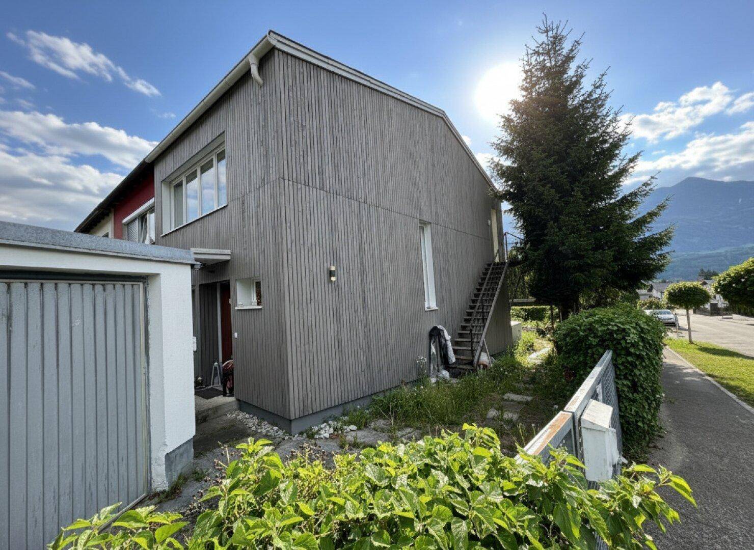 Seitenansicht eines Hauses mit Holzfassade, Außentreppe und einer Garage unter strahlendem Sonnenschein.