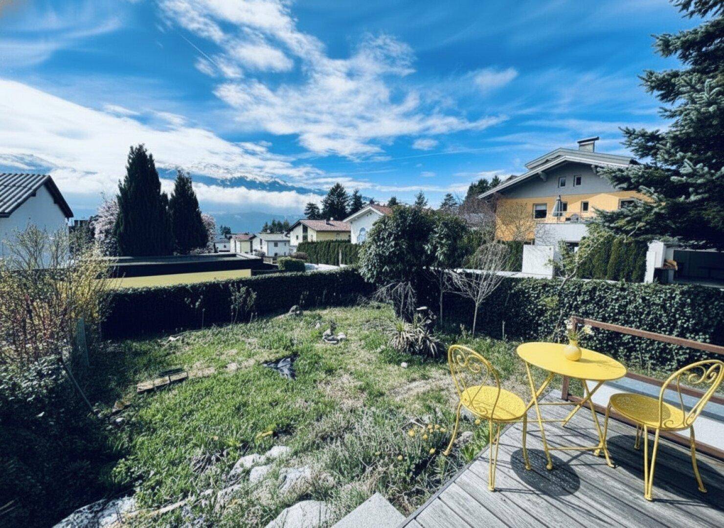 Gartenbereich mit Rasenfläche und einer kleinen gelben Sitzgruppe auf einer Holzterrasse.