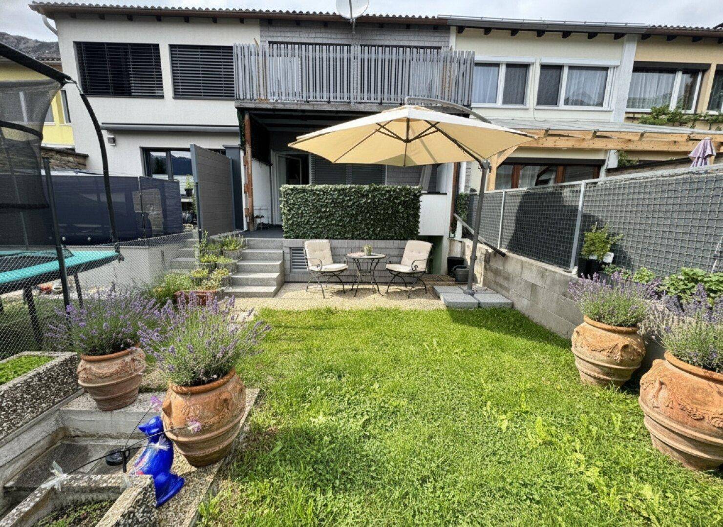 Kleiner Gartenbereich mit Rasenfläche, Blumentöpfen und einer kleinen Terrasse mit Sonnenschirm.