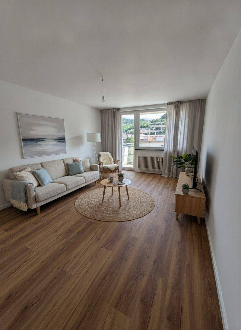 Helles Wohnzimmer mit beigem Sofa, Holzboden und großem Fenster mit Blick ins Grüne.
