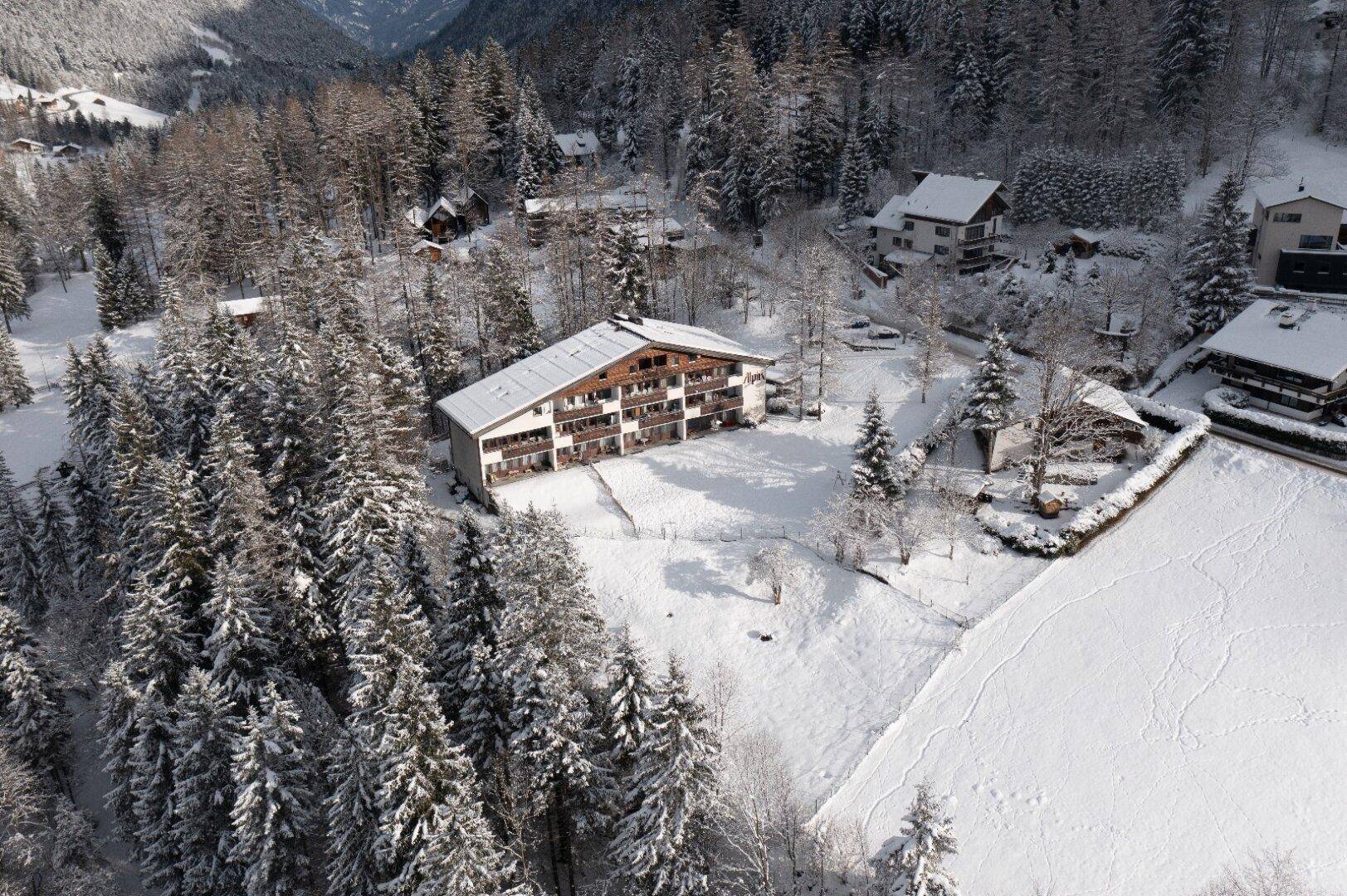 Weitläufige Luftaufnahme eines Gebäudes in einem verschneiten Tal, umgeben von Wald und weiteren Häusern.