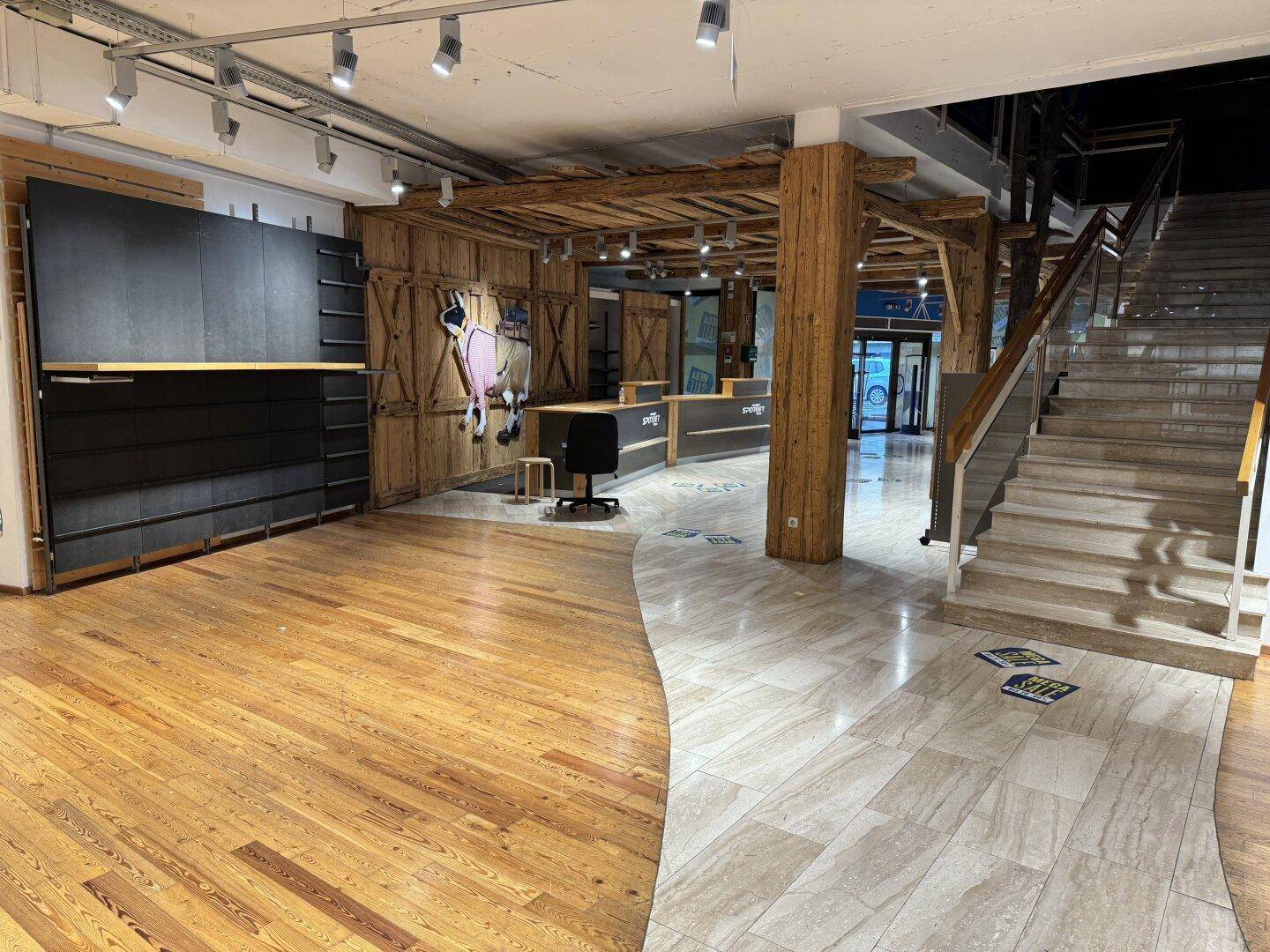 Großer Verkaufsraum mit gemischtem Bodenbelag aus Holz und Fliesen sowie einer Treppe.